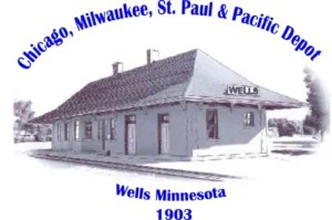 Historic Wells Depot with blue railroad lettering and Wells Minnesota 1903 below.