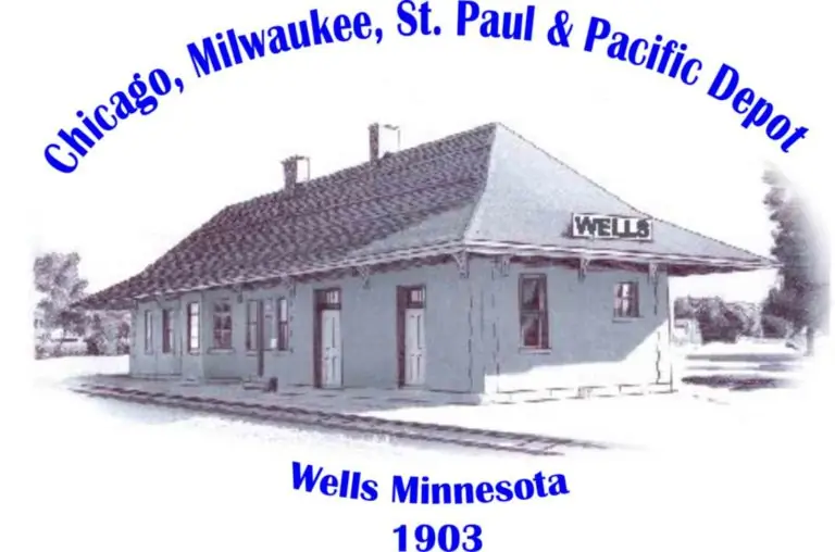 Historic Wells Depot with blue railroad lettering and Wells Minnesota 1903 below.