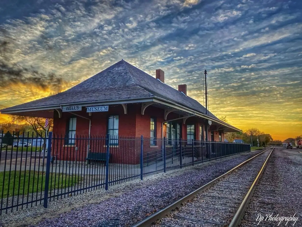 Wells Depot Museum beside railroad tracks under a blue and golden evening sky.