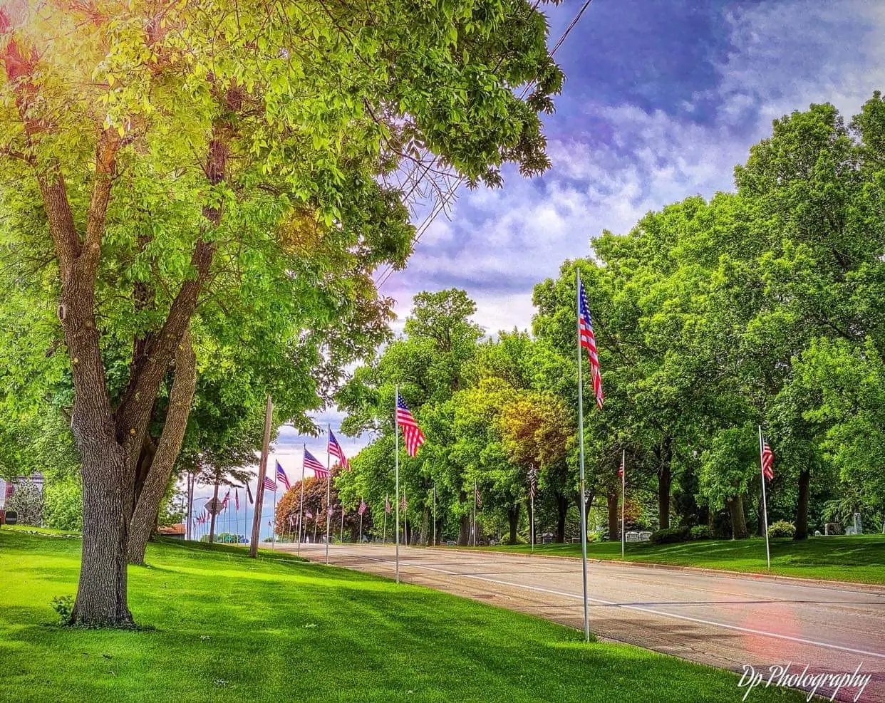 American flags lining a tree-shaded road in Wells beneath a partly cloudy sky.
