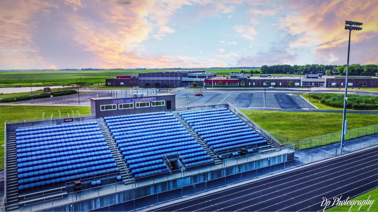 Blue stadium seating and a running track in Wells with school buildings and farmland beyond.
