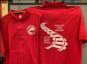 Front and back of a red Wells Depot Museum polo shirt with a white logo and curved railroad track design.